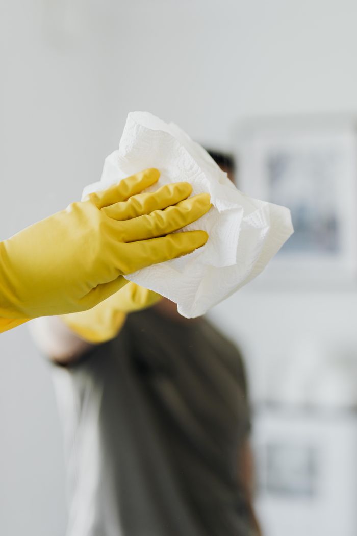about-us-02 Un homme anonyme portant des gants en latex jaunes et un t-shirt sombre nettoie un miroir dans une salle de bain avec une serviette en papier